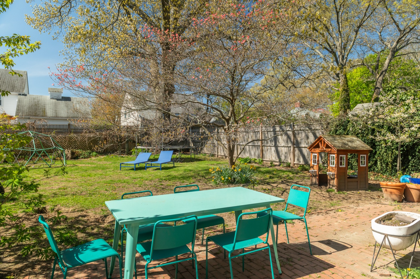 Fenced backyard with a long teal dining table, lounge chairs, and mature trees in bloom.